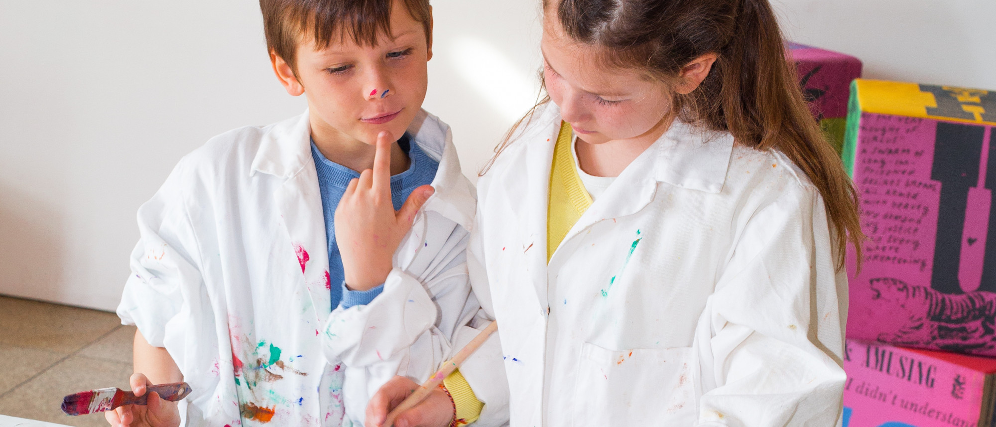 Zwei Kinder mit bekleckertem Mal-Kittel, Farbklecksen im Gesicht und Pinsel in der Hand (MEWO Kunsthalle)